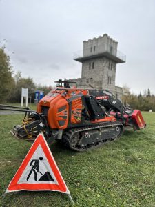 Ein orangefarbenes ferngesteuertes Baufahrzeug und ein dreieckiges „Männer bei der Arbeit“-Schild stehen unter einem bewölkten Himmel auf dem Gras neben einer Mountainbike-Strecke vor einem Steinturmgebäude mit einer Aussichtsplattform.