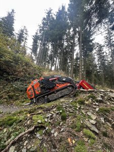 Ein ferngesteuertes Kettenfahrzeug mit der Aufschrift „COMMAND“ fährt einen felsigen, moosigen Hang in einem Wald aus hohen Kiefern hinauf und navigiert durch das zerklüftete Gelände wie ein Mountainbike auf einer anspruchsvollen Strecke.