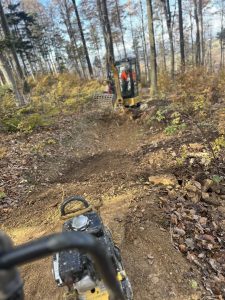 Ein kleiner Bagger arbeitet an einem Feldweg in einem Waldgebiet mit Herbstlaub, während im Vordergrund eine kompakte Rüttelplatte zu sehen ist – perfekt für die Gestaltung dieser neuen MTB-Route für die Mountainbikemovement-Community.