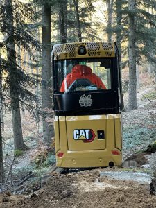 Eine Person in einer roten Jacke bedient einen gelben CAT-Bagger auf einem Feldweg in einem Waldgebiet, umgeben von hohen Bäumen und vereinzeltem Sonnenlicht, und bereitet wahrscheinlich den Weg für Mountainbikemovement-Aktivitäten vor.