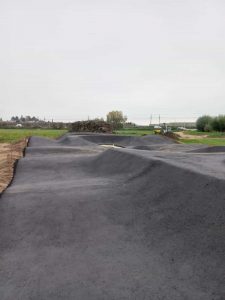 Eine frisch asphaltierte Mountainbike-Strecke mit glattem, dunklem Asphalt und sanften Hügeln liegt in einer offenen Grasfläche unter bewölktem Himmel. Im Hintergrund sind Baumaschinen und Erdhaufen zu sehen.