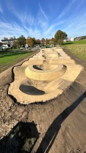 Ein neu angelegter Dirt-Pumptrack mit verschiedenen Steilhängen und Kurven für Mountainbiker, umgeben von Gras und teilweise unbefestigtem Untergrund, unter einem klaren blauen Himmel mit Bäumen und Häusern im Hintergrund.