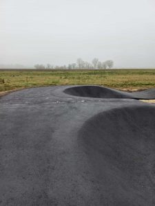 Eine asphaltierte Mountainbike-Strecke mit kurvigen und erhöhten Abschnitten verläuft auf einer offenen Wiese unter einem bewölkten Himmel. In der Ferne sind durch leichten Nebel vereinzelt Bäume zu sehen.