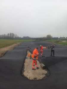 Drei Arbeiter, zwei in leuchtend orangefarbenen Sicherheitsuniformen und einer in dunkler Kleidung, verteilen Kies und glätten Asphalt auf einer großen Außenstrecke, möglicherweise um sie für ein Mountainbike-Event vorzubereiten. Unter dem bewölkten Himmel sind um sie herum Bauwerkzeuge zu sehen.
