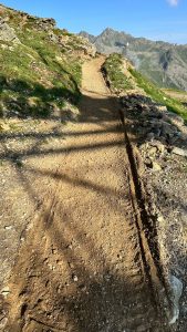 Ein unbefestigter Wanderweg schlängelt sich bergauf durch grasbewachsenes Gelände mit Felsen an den Seiten, die lange Schatten auf den Weg werfen. Im Hintergrund erheben sich schroffe Berge unter einem klaren blauen Himmel.