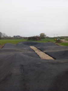 Eine frisch asphaltierte Mountainbike-Strecke mit rollenden Asphalthügeln und einem Mittelstreifen aus freiliegender Erde liegt in einer offenen Grasfläche unter bewölktem Himmel. Im Hintergrund sind Baumaterialien zu sehen.