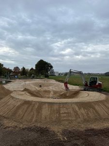Eine Person in Rot bedient einen kleinen Bagger auf einer unbefestigten Baustelle und formt unter einem bewölkten Himmel einen kreisförmigen Erdhügel für eine Mountainbike-Strecke. Im Hintergrund sind Bäume und Gebäude zu sehen.