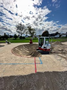 Ein kleiner Bagger formt in einem mit roten und blauen Linien markierten Sandgebiet eine kreisförmige Mountainbike-Strecke; im Hintergrund sind unter einem bewölkten Himmel Kieshaufen und grünes Gras mit Bäumen und Häusern zu sehen.