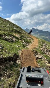 Unter einem teilweise bewölkten Himmel schlängelt sich ein Feldweg einen felsigen Berghang hinauf. Im Vordergrund sind Baumaschinen zu sehen und davor eine hölzerne Rampenkonstruktion – perfekt für das nächste Abenteuer jedes Mountainbike- oder MTB-Fans.