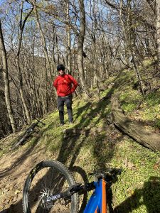 Eine Person in roter Jacke und Helm steht zwischen Bäumen auf einem Mountainbike-Pfad im Wald. Im Vordergrund liegt ein Mountainbike auf dem Boden. Sonnenlicht fällt durch die blattlosen Äste darüber.