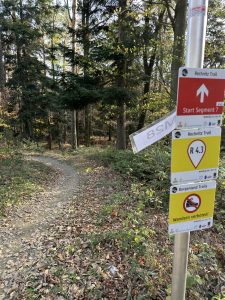 Ein Waldweg schlängelt sich durch herbstliche Bäume. Ein Pfosten am Wegrand zeigt den Rechnitzweg, den Start des Wegabschnitts, die Route R 4.3, und warnt vor der Nutzung bestimmter Wege durch Wandern oder Mountainbiken. Der Boden ist mit Laub bedeckt.