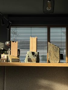 Drei Auszeichnungen aus Holz und Stein stehen auf einem Regal vor einem Fenster mit horizontalen Jalousien. Sonnenlicht wirft Schatten, und darüber hängt eine Lampe. Auf einer der Auszeichnungen steht „BUILDING AWARD 2022“ – eine Auszeichnung für herausragende Leistungen im Wegebau.