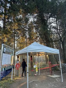 Eine Person steht neben einem weißen Zelt am Eingang einer Mountainbike-Strecke. Schilder, eine Karte und rotes Band markieren den Kontrollpunkt. Im Hintergrund sind hohe Bäume und Sonnenlicht zu sehen.