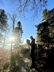 Eine Person steht mit einem Laptop in der Hand auf einem Waldweg, umgeben von hohen Bäumen, durch deren Äste an einem klaren Tag Sonnenlicht fällt. In der Nähe steht ein Mountainbike, das auf eine abenteuerliche Ausfahrt hindeutet.