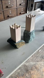 Zwei rechteckige Holzskulpturen mit gezackten, kronenartigen Spitzen stehen aufrecht wie Markierungen auf einem schroffen Pfad, auf rauen Steinen auf einer grauen Tischplatte in einer Werkstatt voller Schubladen und Mountainbike-Bewegungswerkzeugen.