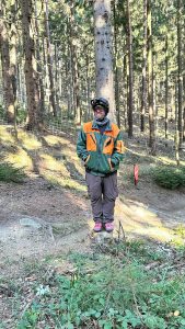 Eine Person in einer grün-orangen Jacke, braunen Hosen, Handschuhen und einem Helm steht auf einer Mountainbike-Strecke in einer sonnenbeschienenen Waldlichtung in der Nähe eines Baumes, im Hintergrund sind hohe Kiefern und grünes Unterholz zu sehen.