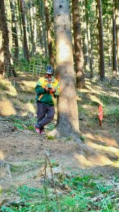 Eine Person in grün-gelber Jacke, Helm und Sonnenbrille lehnt an einem Baum im sonnenbeschienenen Wald und schaut auf ihr Handy – ganz im Mountainbike-Geist. Im Hintergrund sind ein Drahtzaun und eine rote Kreismarkierung zu sehen.