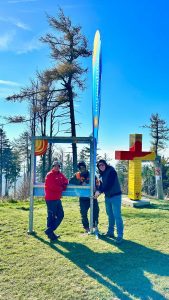 Drei Personen stehen im Freien auf einer Wiese neben einer großen Fachwerkkonstruktion, einer bunten vertikalen Flagge und einem roten Kreuz mit Schildern – eine ideale Kulisse für jeden Mountainbike- oder Trail-Enthusiasten. Kahlköpfige Bäume und ein strahlend blauer Himmel runden die Szene ab.