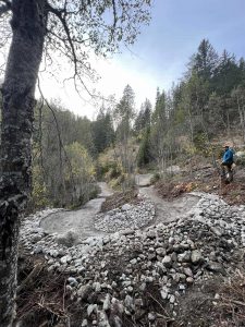 Ein gewundener Feldweg schlängelt sich durch einen felsigen, bewaldeten Hang. Auf der rechten Seite steht eine Person mit einem Rucksack, umgeben von kleinen Bäumen und Steinen unter einem bewölkten Himmel.