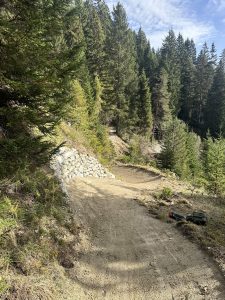 An einem sonnigen Tag schlängelt sich ein unbefestigter Mountainbike-Pfad durch einen Wald mit hohen immergrünen Bäumen. Auf der linken Seite befindet sich eine Stützmauer aus Stein und auf dem Boden liegen einige Werkzeuge – perfekt für jeden Streckenenthusiasten oder Mountainbikemovement-Fan.