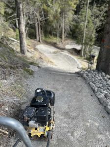 Eine Rüttelplatte steht auf einer Schotterstrecke für Mountainbikes in einem Waldgebiet. Im Hintergrund säumen Bäume und Felsen den gewundenen Weg.