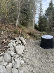 Ein Steinhaufen und ein schwarzer Plastikeimer liegen auf einem unbefestigten Mountainbike-Track in einem Waldgebiet, im Hintergrund Bäume und Grün. Die Szene scheint Teil eines Wanderwegs oder einer Baustelle zu sein.