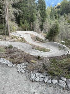 Ein gewundener Schotterweg schlängelt sich durch einen bewaldeten Hügel mit grünen Bäumen auf beiden Seiten und einem teilweise bewölkten Himmel vor Ihnen – das perfekte Gelände für Mountainbike-Abenteuer, mit Felsen entlang der Streckenabschnitte, die für jeden Mountainbike-Enthusiasten eine Herausforderung darstellen.