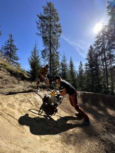 An einem sonnigen Tag schieben zwei Personen eine Schubkarre einen Feldweg in einem Waldgebiet hinauf, mit hohen Kiefern und einer strahlenden Sonne am klaren blauen Himmel.