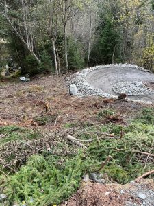 Ein teilweise angelegter, kreisförmiger Schotterweg für Mountainbiker ist von Bäumen und Wald umgeben. Frisch geschnittene Äste und Baumstämme liegen verstreut auf dem geräumten Boden im Vordergrund.