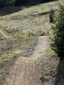 Ein Feldweg an einem grasbewachsenen Hang führt zu einem Baustellen-Warnschild mit einem Arbeitersymbol. In der Nähe steht ein Metallmast, im Hintergrund sind Bäume zu sehen. Das unebene Gelände ist geprägt von losen Steinen und Grasflecken.