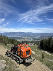 Ein orangefarbener, ferngesteuerter Raupenmäher auf einem Erdhang mit Blick auf ein malerisches Tal, das sich perfekt für Mountainbike-Abenteuer eignet, mit Bäumen, Feldern, einem See und Bergen in der Ferne unter einem blauen Himmel mit dünnen Wolken.