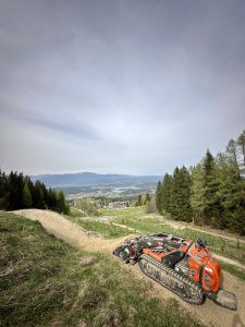 Ein kleiner, orangefarbener, ferngesteuerter Raupenmäher steht auf einem Feldweg an einem grasbewachsenen Hang und überblickt eine Talstadt und einen immergrünen Wald – das perfekte Gelände für Mountainbike-Abenteuer unter dem bewölkten Himmel.