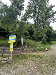 Ein unbefestigter Mountainbike-Trail in einem grünen Waldgebiet ist mit rot-weißem Band abgesperrt. Schilder mit Warnungen und Informationen sind in der Nähe eines Holzzauns aufgestellt, und neben dem abgesperrten Eingang liegt ein dreieckiges Warnschild auf dem Boden.