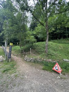 Ein unbefestigter Mountainbike-Pfad durch ein Waldgebiet ist mit rot-weißem Band und einem dreieckigen Baustellenschild abgesperrt, das auf die Sperrung hinweist. Bäume und grünes Gras umgeben die Szene.