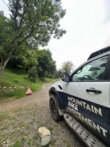 Ein weißer SUV mit der Aufschrift „Mountainbikemovement“ an der Seite steht auf einem Schotterweg in einem Waldstück, wo Absperrband und ein Warnschild die Mountainbike-Strecke vor Ihnen blockieren.