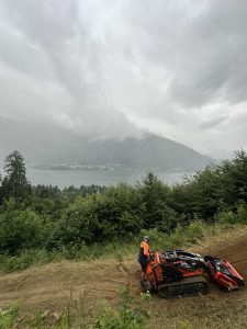 Eine Person in einer orangefarbenen Weste bedient eine kleine Raupenmaschine auf einem Feldweg mit Blick auf einen See und einen nebligen, baumbedeckten Berg unter einem bewölkten Himmel. Dichtes grünes Laub umgibt das Gebiet.