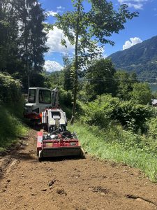 Ein kleiner Bagger und eine rote Kompaktmaschine stehen auf einem Feldweg, umgeben von üppigem Grün, was auf die Arbeit an einem Mountainbike-Trail hindeutet, mit Bäumen, Bergen und einem teilweise bewölkten Himmel im Hintergrund.