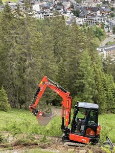 Ein kleiner roter Bagger gräbt einen neuen Weg in einen grasbewachsenen Hang in der Nähe hoher Kiefern. Im Hintergrund über dem Hang ist eine Stadt voller Gebäude zu sehen – eine Szene, die den Geist der Mountainbike-Bewegung einfängt.