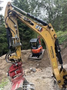 Nahaufnahme eines gelben CAT-Baggerarms mit Hydraulikkomponenten im Fokus; im Hintergrund ist ein weiterer orangefarbener Bagger auf einer Baustelle zu sehen, der einen neuen, von Bäumen umgebenen Weg vorbereitet.