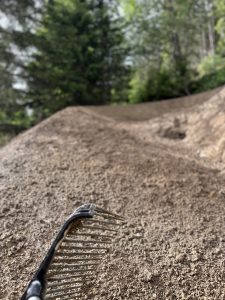 Nahaufnahme eines Metallrechens auf einem unbefestigten Mountainbike-Track mit einem Wald aus grünen Bäumen im Hintergrund. Der Trail führt nach oben, und das Bild hat eine geringe Tiefenschärfe, wodurch der Hintergrund unscharf wird.