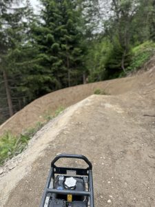 Ein unbefestigter Mountainbike-Trail schlängelt sich durch einen Wald mit immergrünen Bäumen. Im Vordergrund ist ein Baugerät zu sehen, vermutlich zum Verdichten der Wegoberfläche. Die Szene spielt im Freien und ist bewölkt.