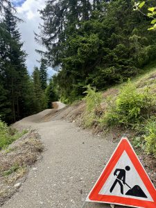 Ein schmaler, gewundener Pfad durch einen Wald. Im Vordergrund steht ein dreieckiges Baustellenschild, das auf bevorstehende Straßenarbeiten hinweist. Hohe grüne Bäume säumen den Weg zu beiden Seiten – perfekt für eine abenteuerliche Mountainbike-Tour.