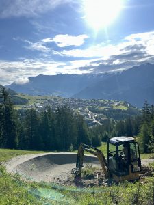 Ein kleiner Bagger arbeitet an einem unbefestigten Mountainbike-Pfad, der von Kiefern umgeben ist. Im Hintergrund sind eine Stadt und Berge zu sehen, unter einem teilweise bewölkten Himmel und hellem Sonnenlicht.