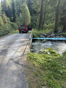Ein roter Pickup mit offener Tür steht auf einer kleinen Brücke über einem fließenden Bach, umgeben von Bäumen und Grün, und markiert den Beginn eines malerischen Mountainbike-Trails in einem Waldgebiet.