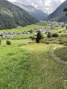 Ein üppig grünes Tal, umgeben von bewaldeten Bergen. In der Mitte liegt unter einem wolkigen Himmel ein kleines Dorf mit weißen Häusern und roten Dächern. Ein gewundener Pfad schlängelt sich durch die grasbewachsenen Hügel und eignet sich perfekt für Mountainbike-Abenteuer ins Dorfzentrum.