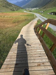 Der Schatten einer Person auf einem hölzernen Promenadenweg in einem grünen Tal, in der Ferne sind unter einem teilweise bewölkten Himmel Berge und ein Dorf zu sehen.