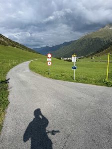 Eine asphaltierte Straße schlängelt sich durch ein grünes Tal mit Bergen im Hintergrund. Entlang der Strecke sind Verkehrsschilder zu sehen, und im Vordergrund erscheint der Schatten einer Person. Der Himmel ist überwiegend bewölkt.