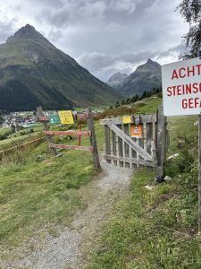 Ein Holztor und Warnschilder stehen am Eingang eines grasbewachsenen Bergpfades, im Hintergrund ein Dorf und dramatische, wolkenbedeckte Gipfel.