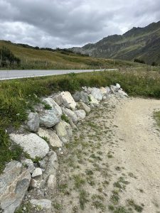 Ein von Felsen gesäumter Schotterweg schlängelt sich neben einem grasbewachsenen Straßenrand entlang, mit grünen Hügeln und Bergen unter einem bewölkten Himmel im Hintergrund – die perfekte Kulisse für ein Mountainbike-Abenteuer.