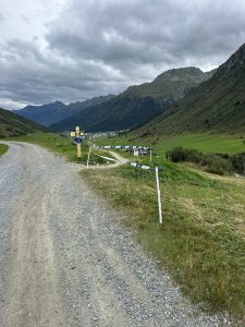 Ein Schotterweg schlängelt sich durch ein grünes Tal mit Bergen im Hintergrund und bewölktem Himmel. Gelbe Wegweiser und eine weiße Absperrung markieren eine Weggabelung – ideal für Mountainbiker, die in ein entferntes Dorf fahren.